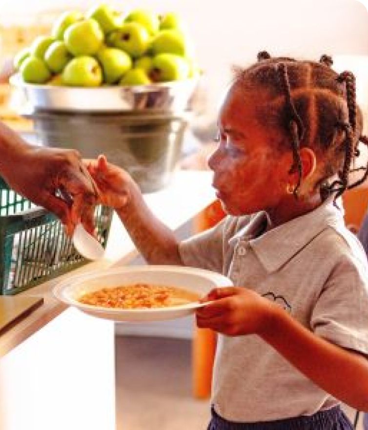 A small child, wearing school uniform, being given food, holding their plate out for an adult dishing up for them and accepting an eating utensil. Part of the projects for us Giving Back