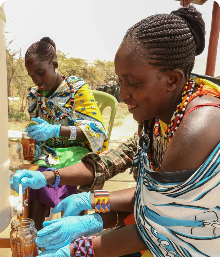 2 Ladies, wearing traditional attire, bottling honey with smiles on their faces. This image also found on our homepage.