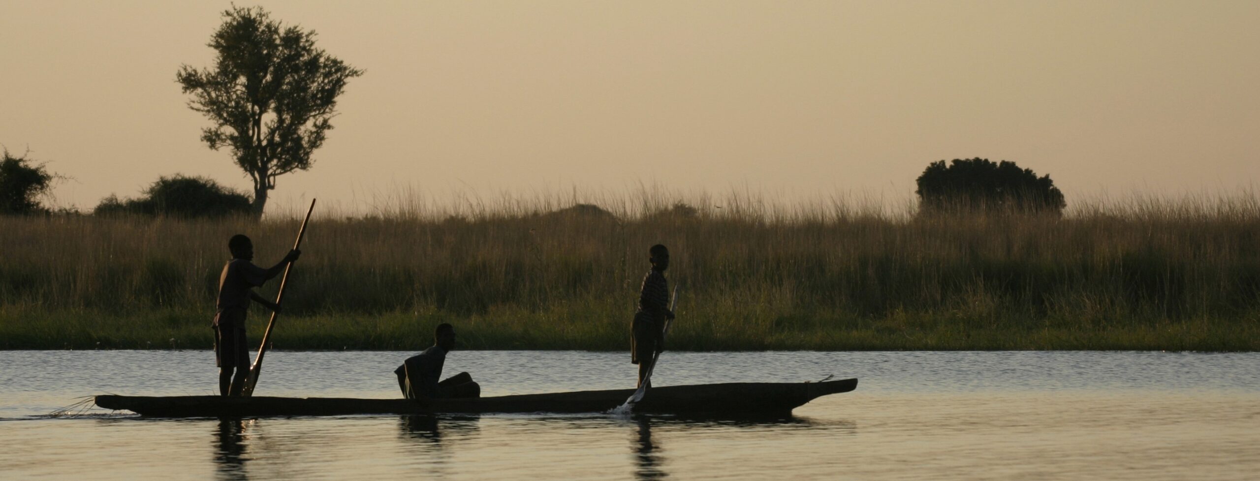 A mokoro at dusk as one of the highlights of Chobe floating across the water carrying 3 people and reeds in the background.