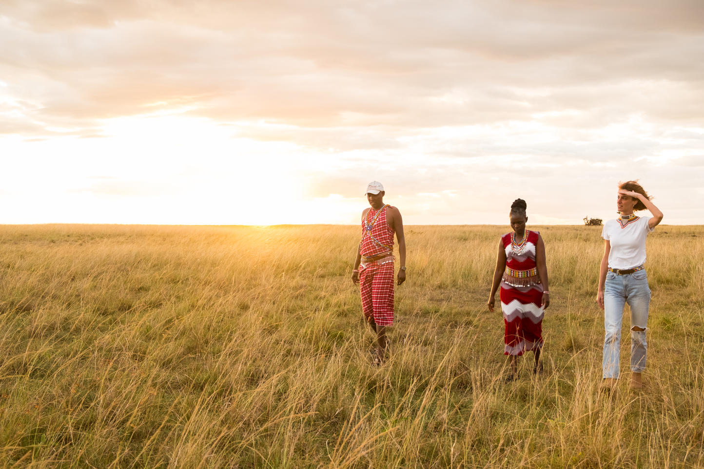 2 local villagers and a tourist walking together in the Savannah with the sun setting behind them.