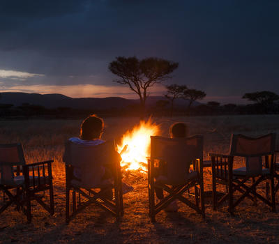 Campers sitting at a roaring fire with the Savannah in the back ground with silhouetted Acacia Trees in Ndutu