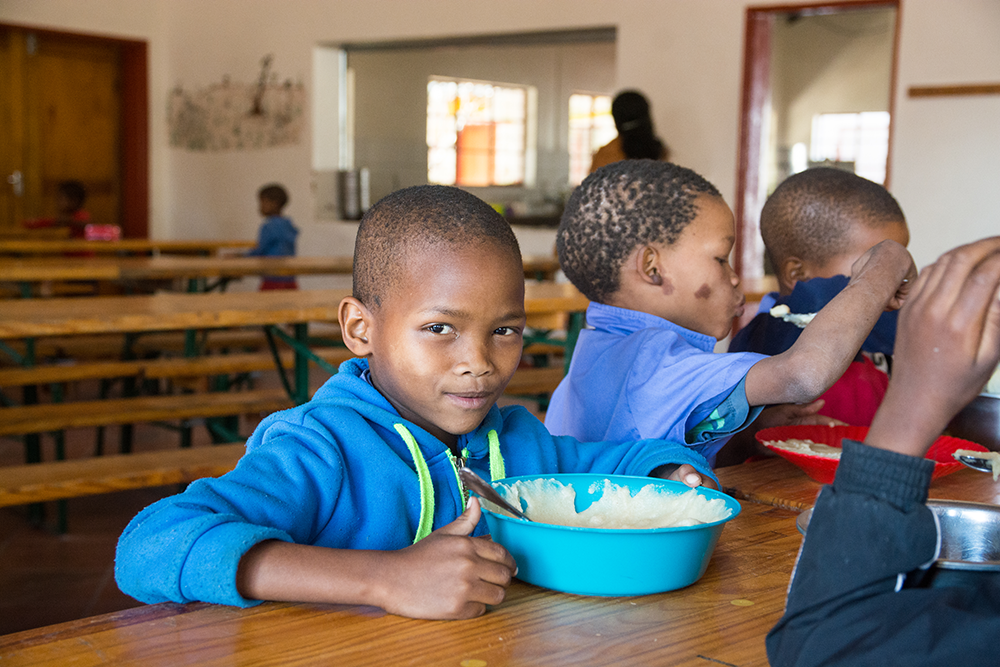 Children with big smiles, sitting down for a meal in support from Mama Africa Foundation.