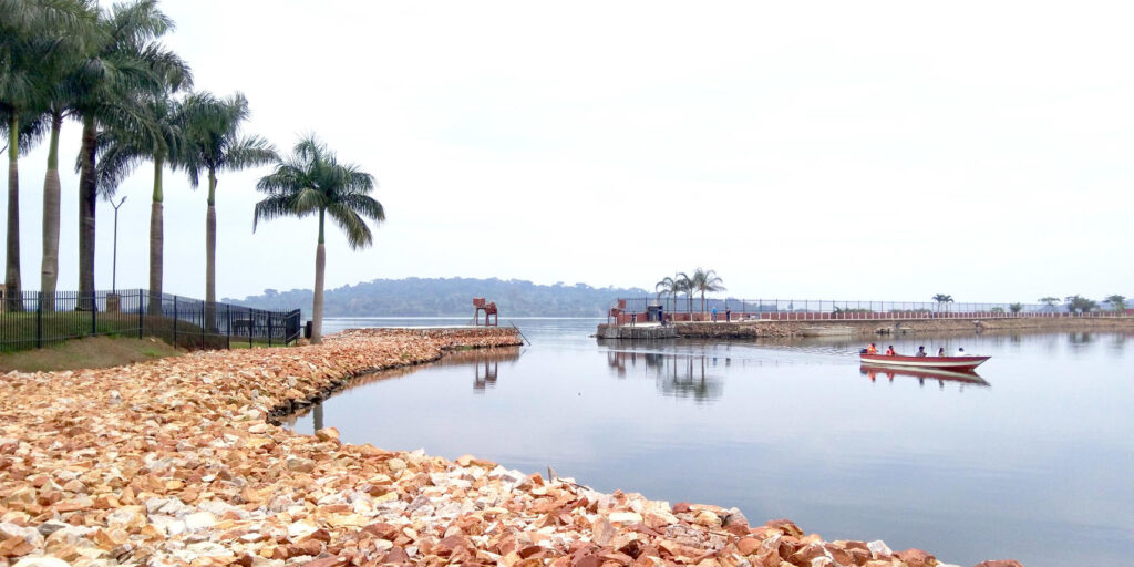 Palm trees on the left over a pebble beach by the lake. Boats on the lake and a bridge is visible.