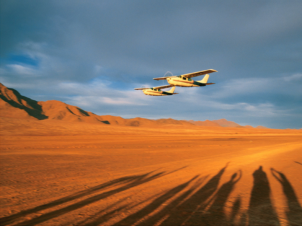 2 planes flying low side by side under an overcast sky which contrasts the red sand below. silhouettes of guests in the sand show them welcoming the planes and some taking photos.