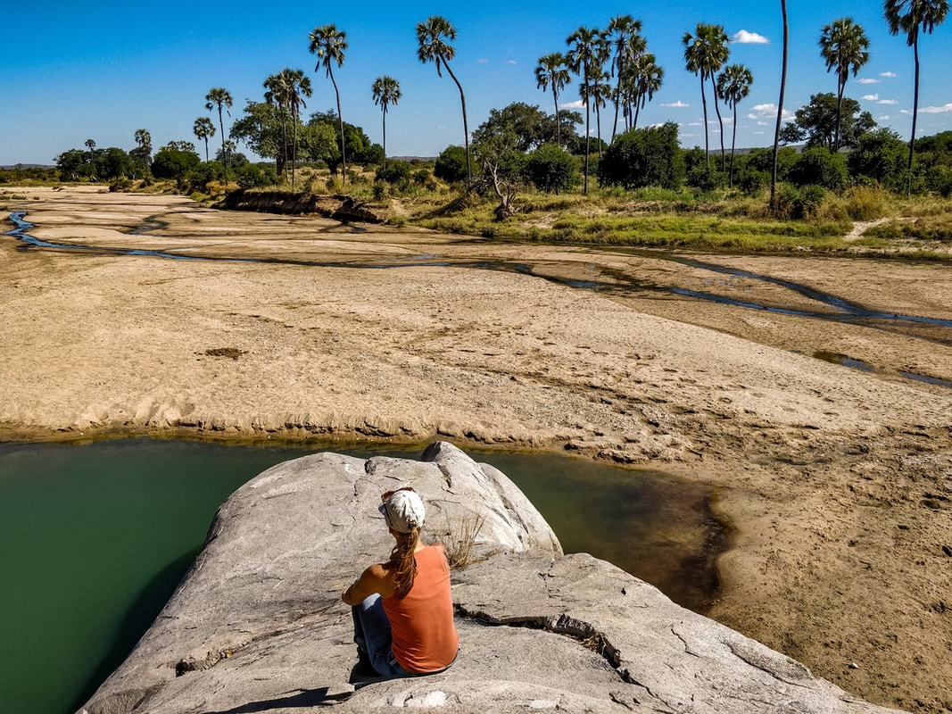 A guest enjoying herself on a rock by herself at the water hole. Elephant prints can be seen in the sand on the other side of the water.