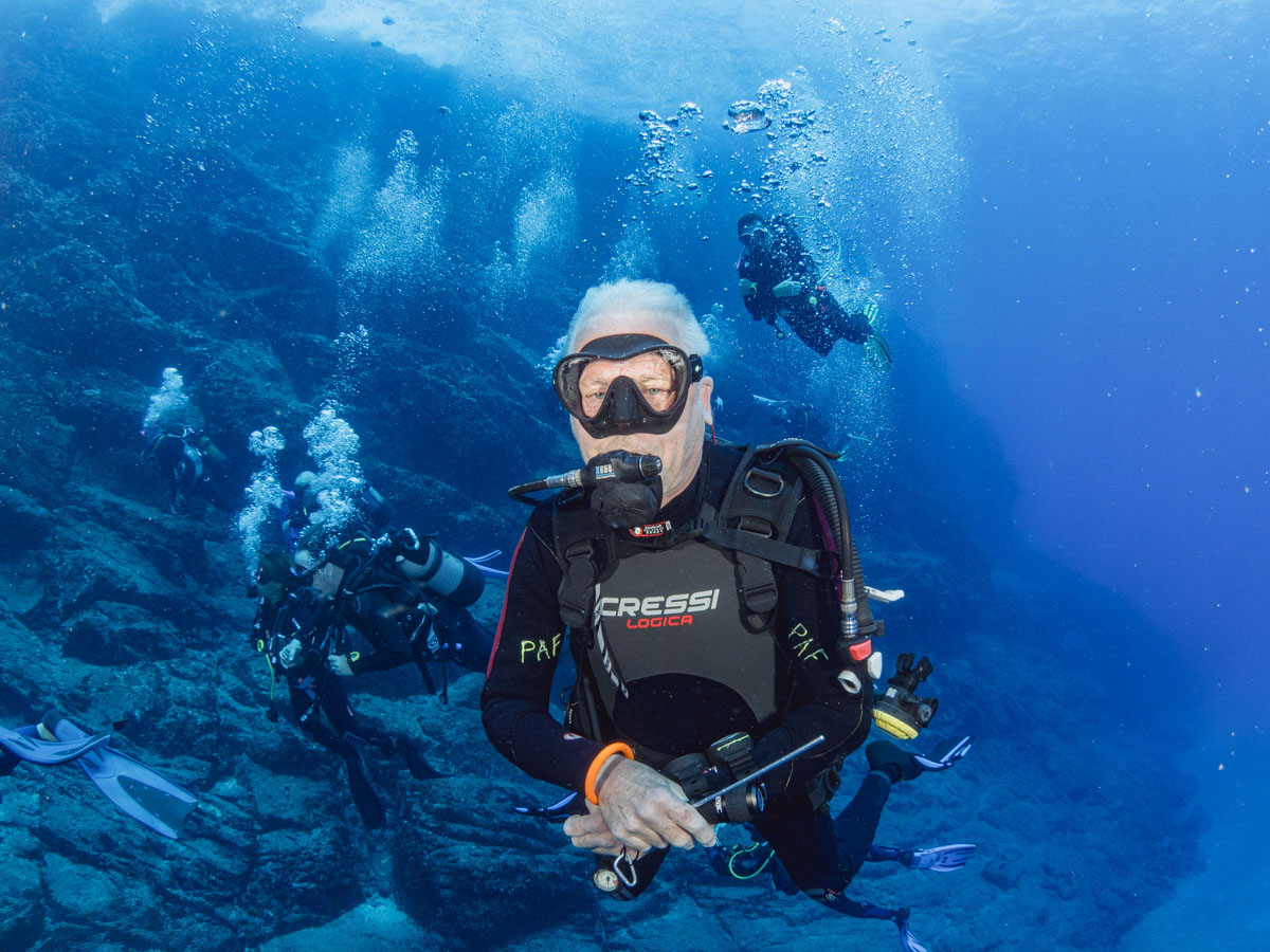 A scuba diver in St Helena looking at the camera. Behind him are other scuba divers diving in the crystal clear water along some rocky reef.